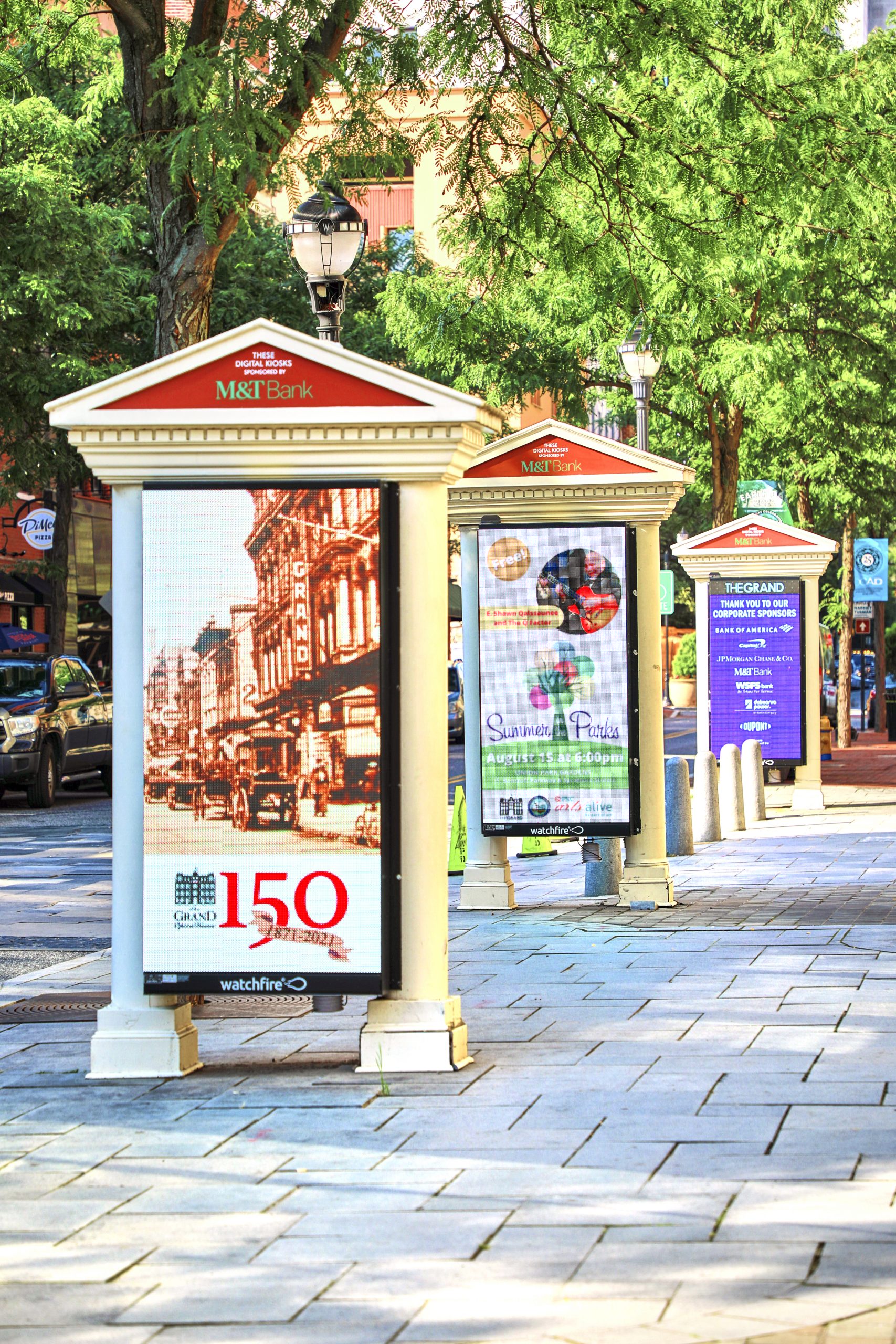 Three tall digital message boards fitted in front of a building on a stone walkway area